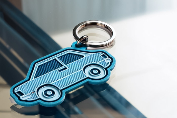 Schlüsselanhänger aus PVC in Autoform, blau mit geprägter Struktur, auf Glasoberfläche liegend.