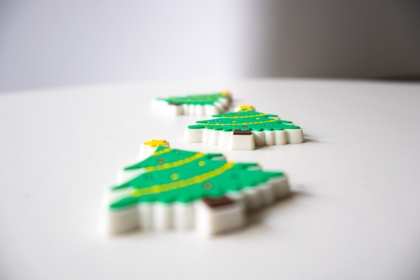 Drei weihnachtliche Gummi-Magnete in Form geschmückter Tannenbäume liegen auf einer weißen Oberfläche, mit unscharfem Hintergrund.